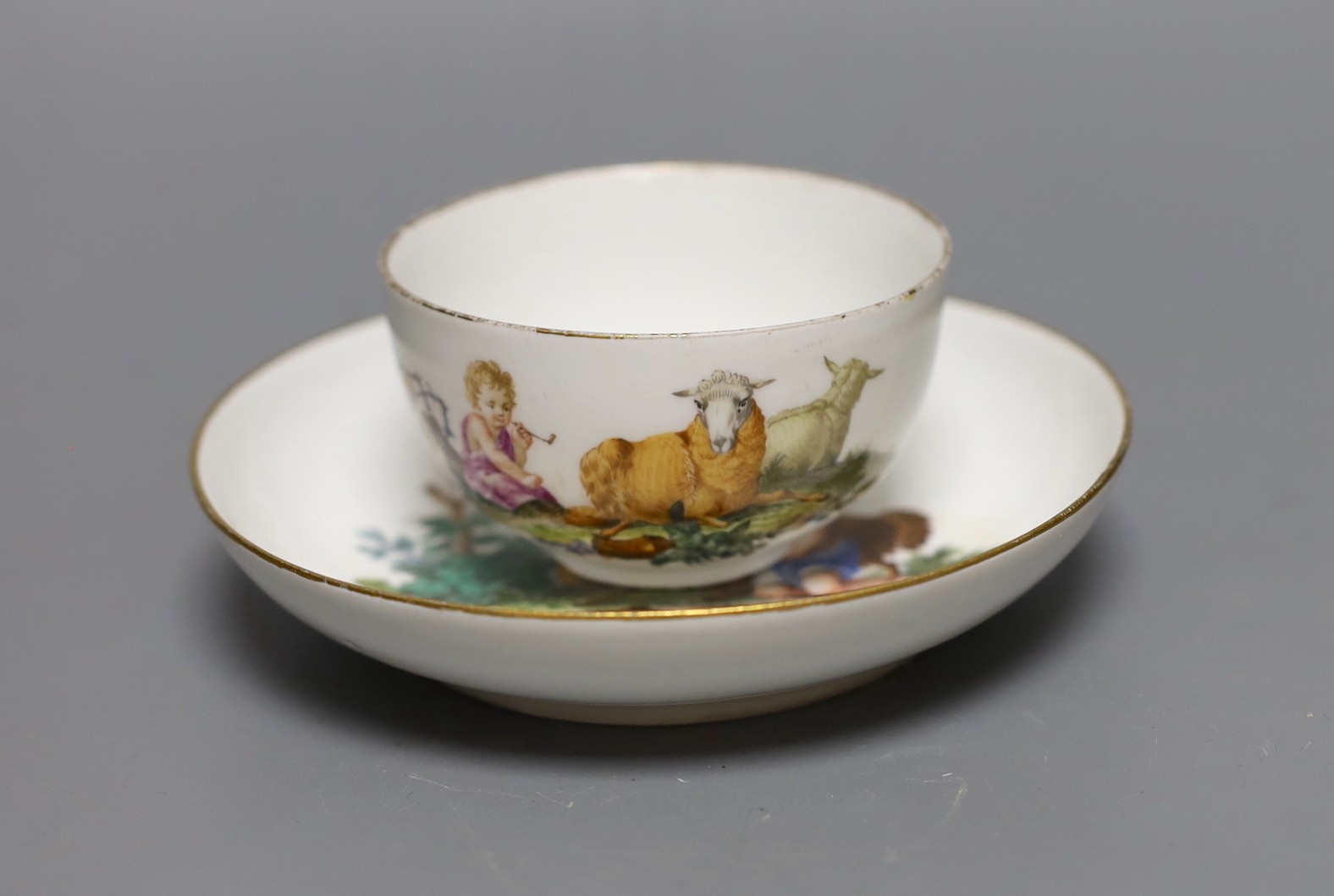 A Meissen teacup and saucer painted with a landscape with children in robes with recumbent sheep, crossed swords mark and dot in underglaze-blue, impressed numerals, saucer 13.5 cms diameter